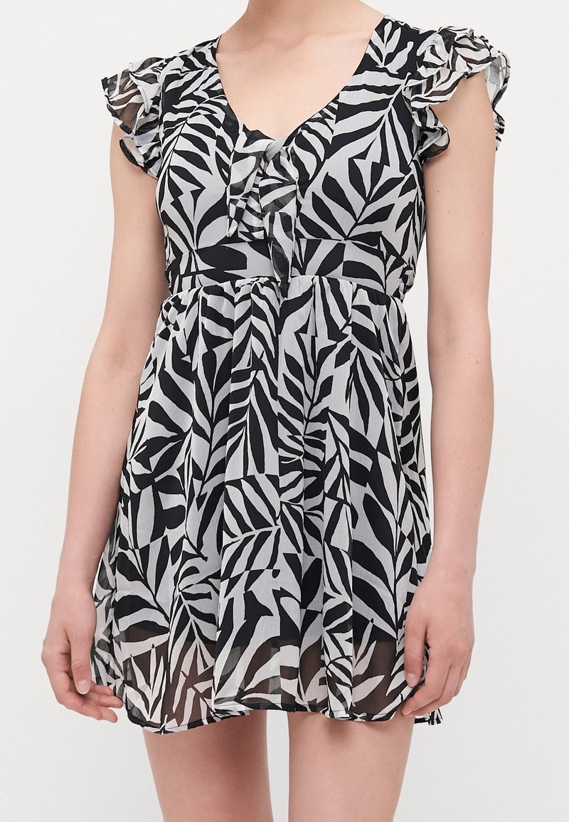 Femme portant une robe sans manches noire et blanche à motif de feuilles, avec des détails à volants sur les épaules et un décolleté en V.