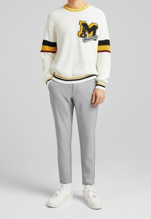 Mann trägt weißen Strickpullover mit schwarzen, gelben und roten Streifen sowie graue Hose, steht mit einer Hand in der Tasche und weißen Turnschuhen.