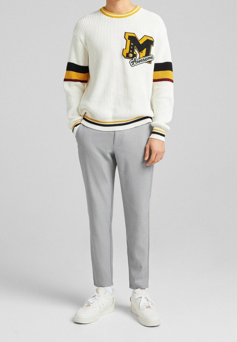 Homme portant un pull en tricot blanc avec des rayures noires, jaunes et rouges, et un pantalon gris, debout avec une main dans la poche et des baskets blanches.