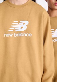 Smeđi dukserica od mekanog materijala s bijelim "New Balance" logotipom. Oversized kroj s spuštenim ramenima i klasičnim okruglim izrezom.
