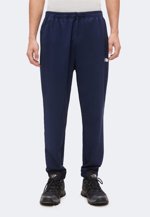Homme portant un pantalon de sport bleu marine et des chaussures de randonnée noires avec des logos de marque visibles sur le pantalon et les chaussures, debout devant un fond blanc.