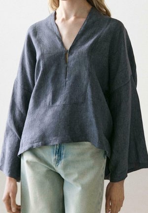 Femme portant un chemisier ample gris foncé à col en V et manches longues, ainsi qu’un jean bleu clair, montrée de la poitrine aux hanches sur un fond uni.