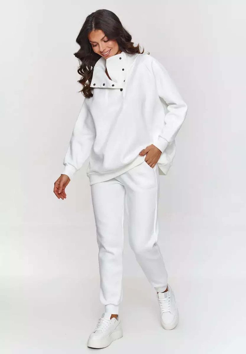 Biała, luźna bluza z wysokim kołnierzem i detalami na zatrzaski, połączona z białymi spodniami dresowymi i białymi sneakersami. Gładka tekstura.