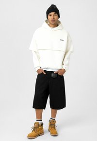Witte oversized hoodie met gelaagd ontwerp, zwart logo, zwarte shorts, gele laarzen en een zwarte muts. Gestructureerde stof en zichtbare hardware.