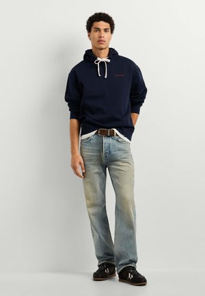 Junger Mann mit lockigem Haar, der einen dunkelblauen Hoodie, hellblaue Jeans und schwarze Sneakers trägt, steht vor einem schlichten weißen Hintergrund.