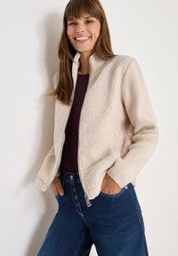 Beige bomber jacket with a soft, textured fleece front and ribbed long sleeves. Features a front zipper closure and side pockets, paired with dark blue jeans.