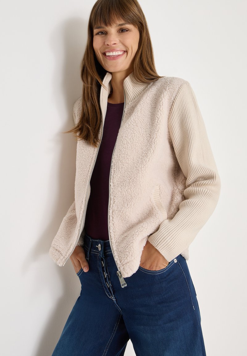 Giacca bomber beige con morbido pannello in pile testurizzato nella parte anteriore e maniche lunghe a costine. Chiusura frontale con zip e tasche laterali, indossata con jeans blu scuro.