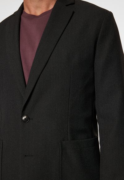 Blazer nero in tessuto texturizzato con chiusura a un bottone e due tasche laterali. Indossato sopra una t-shirt a girocollo bordeaux.