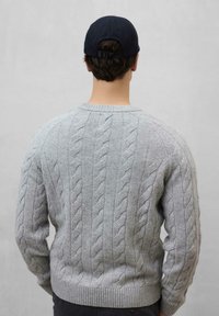 Suéter gris de punto con trenzas y ribetes acanalados, hecho de una tela suave. Usado con una gorra oscura, visto desde atrás.