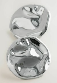 Due dischi metallici lisci, di forma irregolare, con una finitura cromata lucida che riflette la luce, mostrando una texture elegante e lucida.