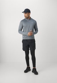 Haut de sport à manches longues gris avec demi-fermeture éclair, shorts noirs, collants noirs et chaussures de course. Casquette noire avec logo. Détails de couture contrastés.