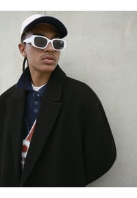 Une jeune personne portant des lunettes de soleil blanches, une casquette blanche, un manteau noir et un pull à motifs bleu marine, se tenant contre un mur gris clair uni.