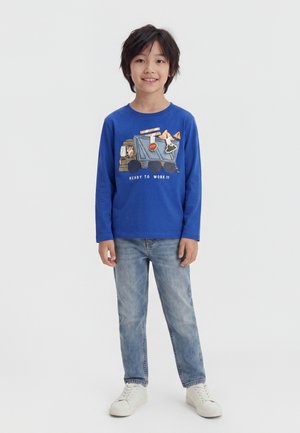 Garçon aux cheveux foncés portant un t-shirt bleu à manches longues avec un motif de camion et un jean, debout et souriant sur un fond blanc.