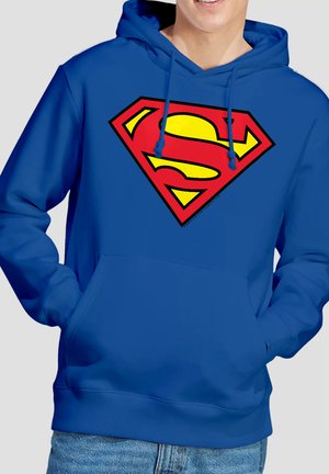 Person trägt einen blauen Kapuzenpullover mit einem großen roten und gelben Superman-Logo auf der Brust, die Hände in der Vorderentasche, lächelt leicht.