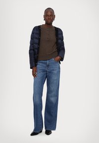 Puffer jacket in navy, brown henley shirt, and light blue wide-leg jeans. Model wears black pointed-toe shoes; hands in pockets.