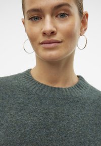 Zelený vlněný svetr s kulatým výstřihem a žebrovanými manžetami. Modelka má na sobě kruhové náušnice, hladkou pleť a jemný make-up, na bílém pozadí.