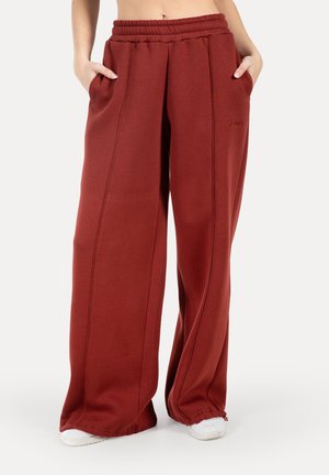 Pantaloni felpati a gamba larga, di colore marrone rossastro, con un elastico in vita, tasche laterali e dettagli di cucitura discreti, realizzati in tessuto morbido.