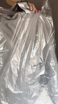 Îmbrăcăminte gri învelită în plastic transparent, suspendată pe un umeraș negru. Textura pare netedă, cu detalii minime vizibile prin folie.