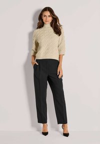 Eine Frau steht vor einem schlichten Hintergrund und trägt einen beigen, strukturierten Rollkragenpullover, schwarze maßgeschneiderte Hosen und schwarze spitz zulaufende High Heels.