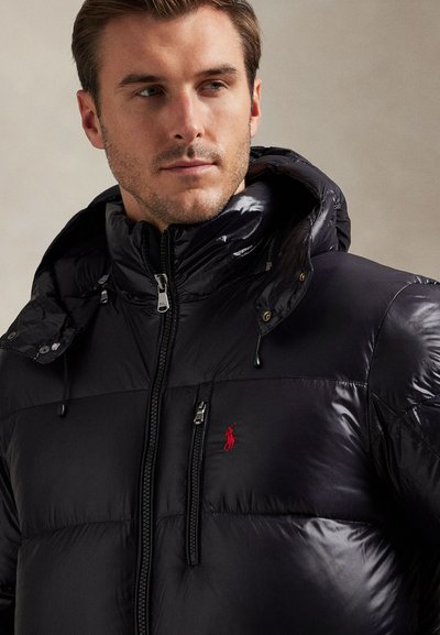 Man wearing black puffer jacket with hood, front zipper, chest pocket, and red embroidered logo, looking slightly to the side.