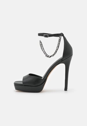 Czarny sandał na wysokim obcasie z platformą, wykonany z teksturowanego materiału syntetycznego, z paskiem wokół kostki zapinanym na klamrę i ozdobą w postaci łańcuszka.