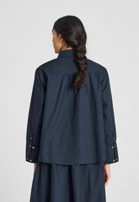 Camicia in cotone blu navy con colletto a bottone, maniche lunghe e design con pieghe sul retro. Presenta dettagli a bottone sui polsini.