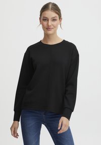 Schwarzes Rundhals Sweatshirt mit langen Ärmeln und lockerem Schnitt. Glatte Textur und sichtbare Nahtdetails. Kombiniert mit blauen Jeans.