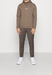 Hombre con sudadera marrón con el texto "LES DEUX", pantalones marrones a juego y zapatillas blancas, de pie contra un fondo claro y liso.