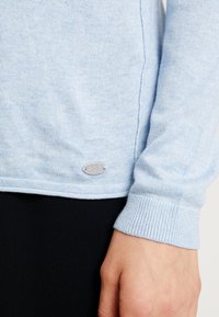 Maglione lavorato a maglia azzurro chiaro con polsini e orlo a coste, caratterizzato da un piccolo logo in argento sul lato vicino all'orlo. Texture morbida.