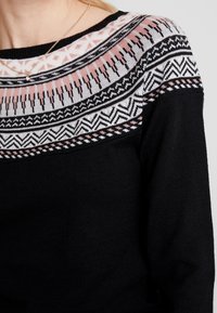 Svart tröja med en färgglad, mönstrad axel i rosa, vitt och svart. Stickad struktur med geometriska och abstrakta mönster.