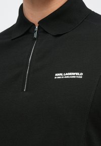 Črna polo majica z ovratnikom na zadrgo, izdelana iz gladkega blaga, z belim logotipom na levi strani prsnega koša.