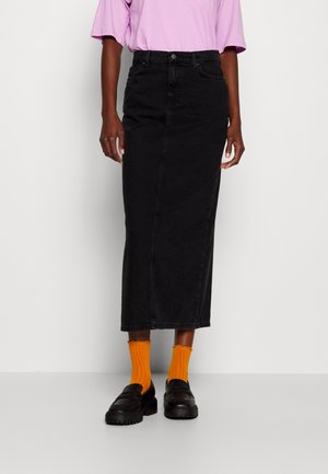 Frau trägt ein helllila kurzärmeliges Shirt, einen hoch taillierten schwarzen Midirock, orangefarbene Socken und schwarze Loafers, steht vor einer weißen Wand.