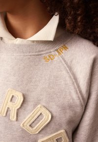 Detailný záber osoby v svetlo béžovej mikine s výšivkou "SD-JPN" v žltej farbe a bielou košeľou pod ňou.