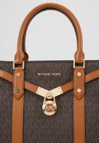 Sac à main en cuir marron avec un motif monogramme, des accents beiges, des accessoires dorés, un détail de verrou et des poignées rondes doubles.