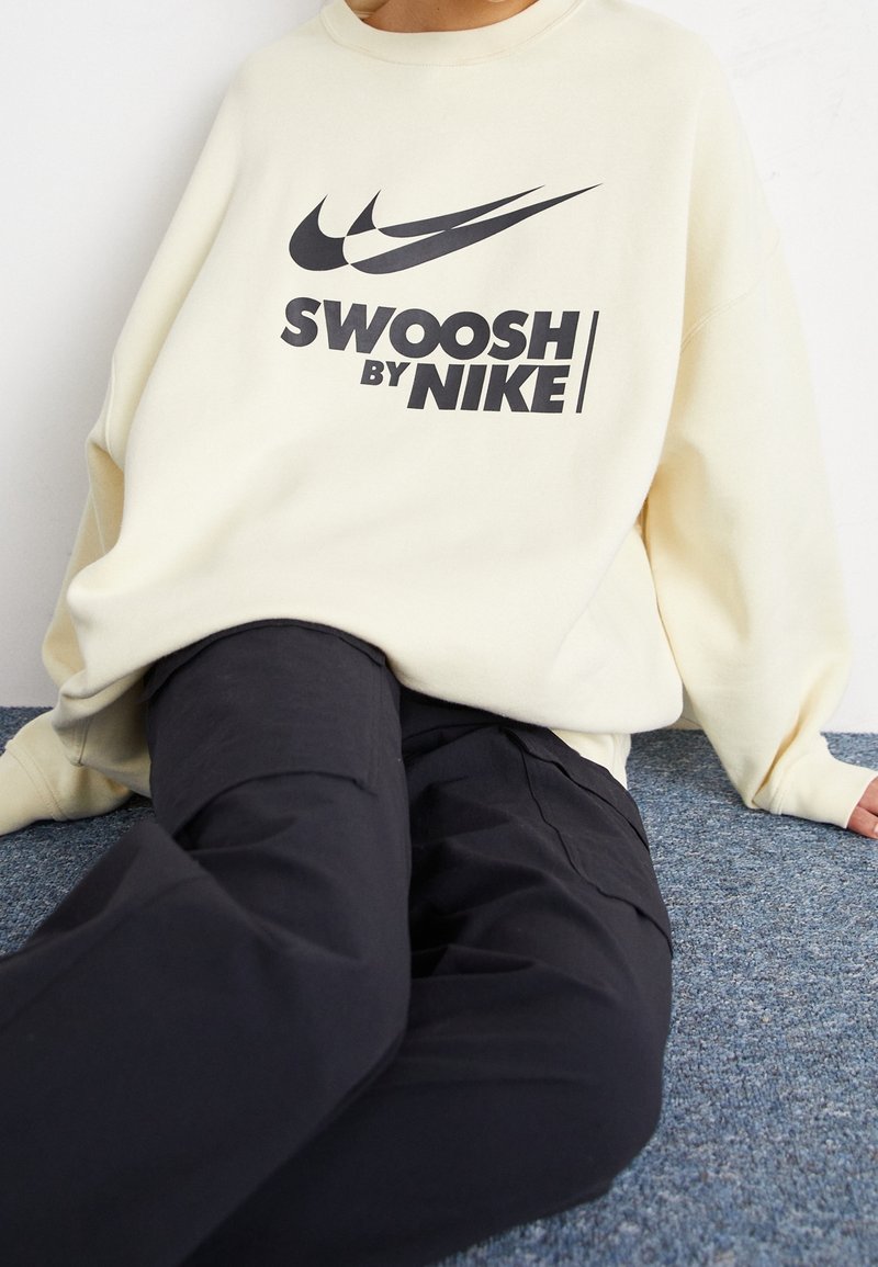 Sudadera color crema con un logo negro "Swoosh de Nike", combinada con pantalones negros. La tela parece suave con un ajuste relajado.