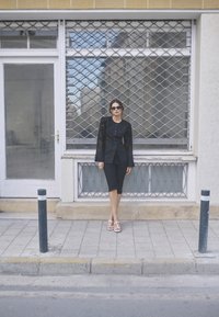 Blazer nero su misura con maniche lunghe trasparenti e bottoni, abbinato a pantaloni corti neri di lunghezza al ginocchio e sandali bianchi; sfondo urbano.