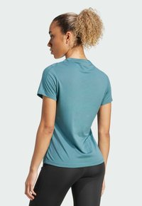 Camiseta deportiva de manga corta color azul verdoso con cuello redondo y textura lisa, combinada con leggings negros. Vista desde atrás.