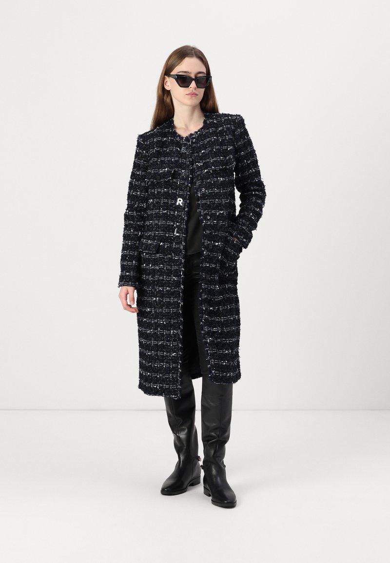 Marineblauer Tweedmantel mit strukturiertem Finish, langen Ärmeln und Fronttaschen. Kombiniert mit enganliegenden schwarzen Hosen und knielangen schwarzen Stiefeln.
