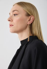 Gold drop earrings feature long, textured chains. The model wears a high-collared black top, showing a sleek, modern design.