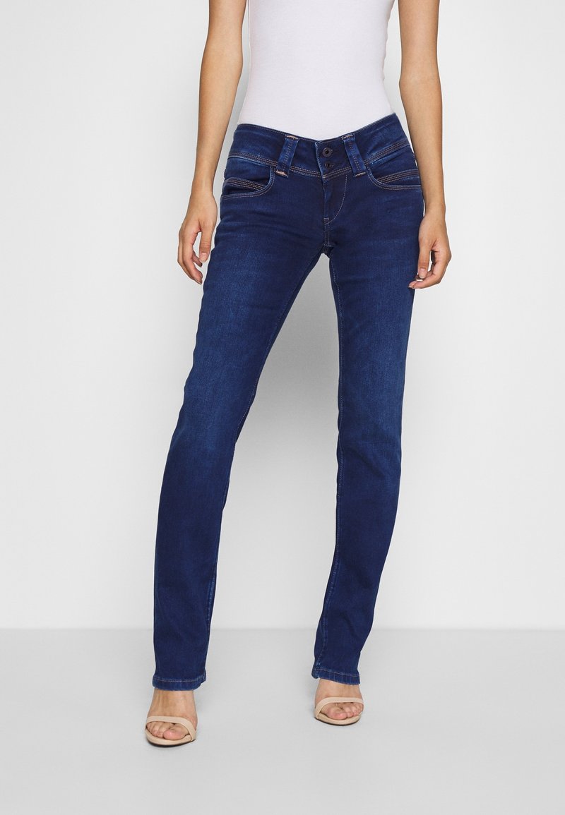 Jeans de mezclilla azul oscuro con corte ajustado, diseño de cinco bolsillos y detalles de costura sutiles. Combinados con una camiseta sin mangas clara y tacones beige.