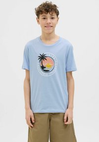 Vaaleansininen puuvillapaita, jossa on pyöreä kuvio, jossa on palmu, auringonlasku ja teksti. Lyhyet hihat, pyöreä kaula-auko ja sileä pinta.