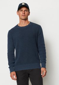 Navyblauer Strickpullover mit strukturiertem Muster, gerippten Bündchen und Saum. Getragen mit einer dunklen Mütze, die ein Logo-Patch aufweist.