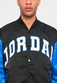 Sort og blå bomberjakke med stort hvidt "JORDAN" tekst, blank overflade, trykknaplukninger og ribbet krave.