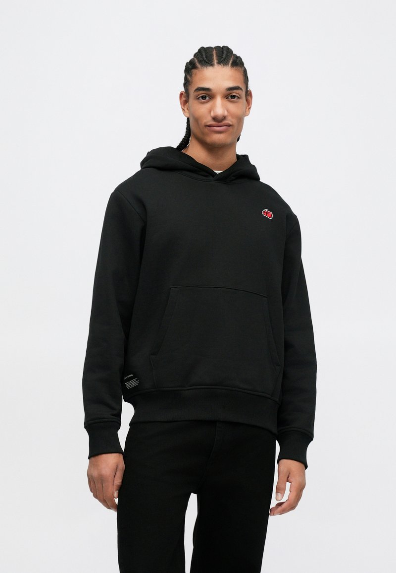 Jeune homme avec des cheveux tressés portant un sweat à capuche noir avec une poche frontale et un petit logo rouge, se tenant devant un fond clair.