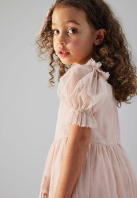 Helles rosa Kleid mit puffärmel, Rüschendetails und strukturierter Rock. Lockige Haare, teilweise mit einer Schleife zurückgesteckt.