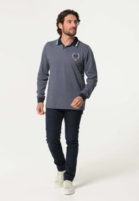 Langarm polo in donker grijs heather, met een geribde kraag met blauwe accenten en een geborduurd logo op de borst, gecombineerd met donkere spijkerbroek.