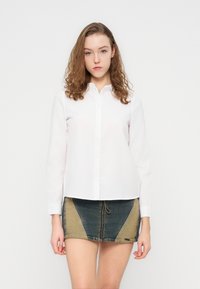 Λευκό πουκάμισο με μακριά μανίκια και κουμπιά, με μυτερό γιακά, συνδυασμένο με μίνι φούστα από τζιν με πάνελ σε μπλε και καφέ αποχρώσεις.