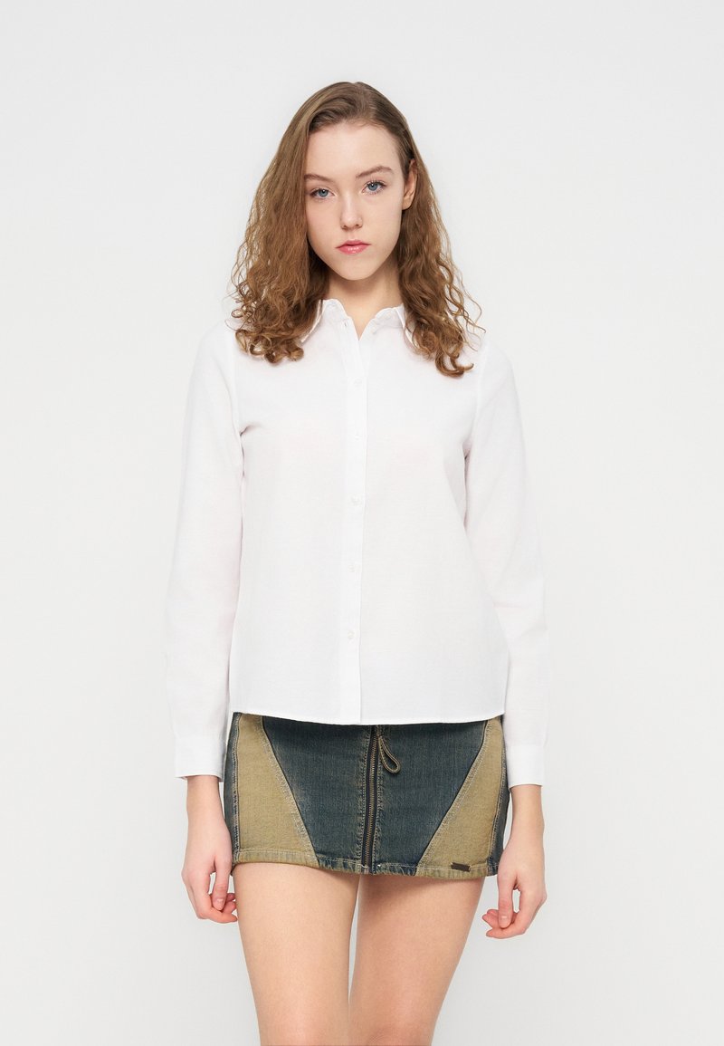 Λευκό πουκάμισο με μακριά μανίκια και κουμπιά, με μυτερό γιακά, συνδυασμένο με μίνι φούστα από τζιν με πάνελ σε μπλε και καφέ αποχρώσεις.