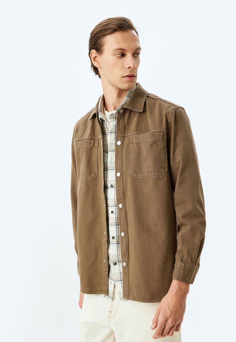 Brun knappad skjorta med två bröstfickor, en kragig hals och en mjuk tygstruktur. Lager på en svart och vit rutig skjorta.