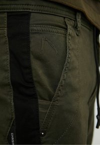 Olivgrön byxor med svarta accenter, en texturerad yta, synlig sömnad och sidfickor. Inkluderar en detalj med tryckknappsstängning.